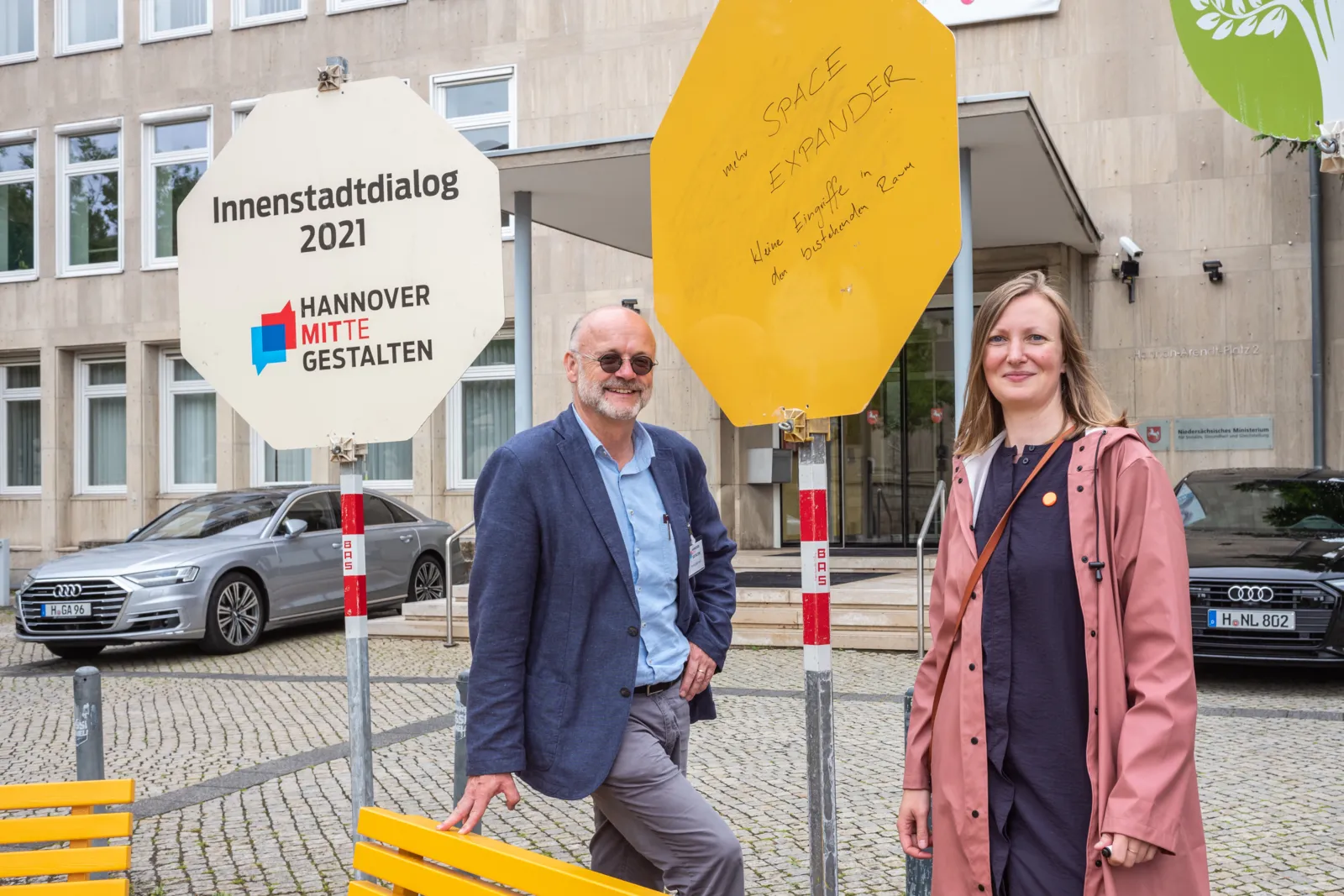 Zwei lächelnde Person an einer Bank vor einem Gebäude.